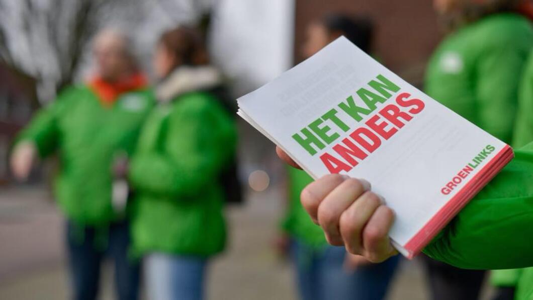 Iemand houdt GroenLinks-flyers vast met daarop: "Het kan anders". De persoon draagt een groen jasje. Op de achtergrond staan meer mensen in groene jasjes.